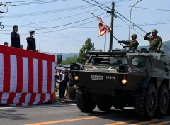 紅白の幕のステージ上に立つ人に向かって、敬礼しながら路上を進む装甲車の写真