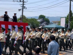 紅白の幕のステージ上に立つ人に向かって敬礼しながら、隊列を組んで歩く自衛官の写真