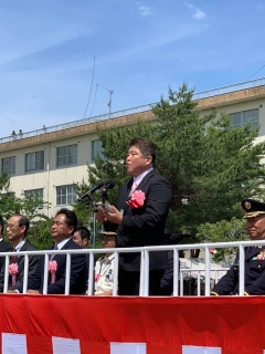 紅白の幕のステージ上に、胸章を着けて座っている自衛官やスーツ姿の関係者と胸章を胸に着けてマイクの前に立って話をしている市長の写真