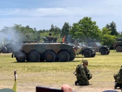 芝生の上で迷彩服をを着た自衛官がしゃがみ、3台の装甲車の後部から煙出ている式典の様子の写真