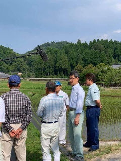 田んぼの淵に立って水の張られた田んぼの方を見ながら立っている河野知事、市長と農家の男性の写真