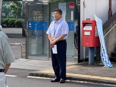 自動ドアのある建物の隣に赤いポストが設置されている路上で半袖シャツの市長が立って話をしている写真