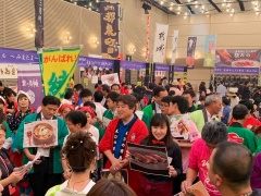 のぼり旗などで飾られた会場内に緑の半被や赤い半被を着た参加者が料理の写真パネルを持っており、周囲に大勢の来場者が集まりにぎわっている様子の写真