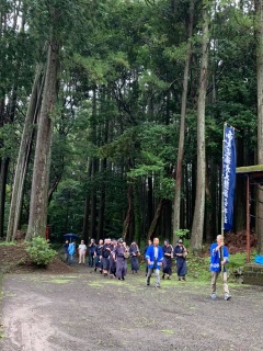 林の奥から歩いてくる、のぼり旗をもった青い半被を着た男性を先頭に、2列になって歩いている祭りの参加者の写真
