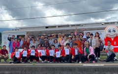 駅のホームに「吉都線大好き！」の文字が書かれたカードを持っている赤い半被をきた学生、その後ろにマスコットキャラクターのみなほと大勢の参加者が集まっている集合写真