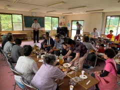 ピンクのカーペットが敷かれた室内で黒板前に立っている半袖のシャツの男性、3列に並べられた料理のおいてあるテーブルの周りに座っている参加者の写真