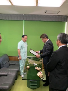 緑のロールカーテンが下ろされた部屋で額縁に入った感謝状を読み上げている市長と、向かい合って立っている林田先生の写真