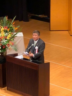 ステージ上の演壇で祝辞を述べている、胸に白い花をつけた市長の写真