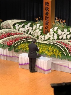 ステージ上に設置された、「戦没者慰霊の碑」がたてられた花祭壇の前でスピーチをしている男性の写真