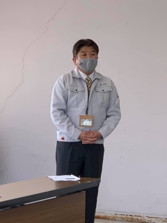 長机の傍でマスクをつけて立っている作業着を着用した市長の写真