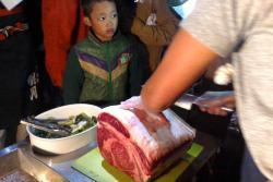 大きな肉の固まりを包丁で切る様子と見物に来た少年が写っている写真