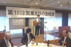 第18回関東えびの会総会と幕がかけられた舞台上にギターを肩からさげた男性が立っている写真
