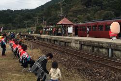 線路に赤い電車が停まっていて、赤い法被を着た人たちが椅子に座って電車を見ている写真