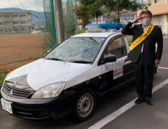 駐車場に停車してあるパトカーの隣で、スーツを着て黄色のタスキをかけ、敬礼している市長の写真