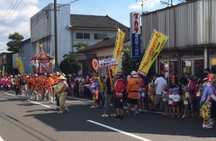 道路を神輿を担いだ人達、のぼり旗を持った人たちが縦に並んで歩いている写真