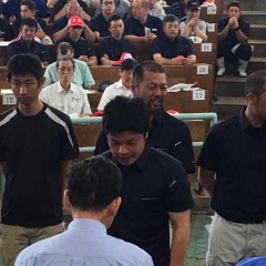 黒い服を着た一人の男性が前に出て水色シャツを着た男性と向かい合わせに立っている写真