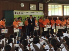着席する子供たちの前に立つ市長と、後方に立つオレンジ色のシャツを着た関係者の写真