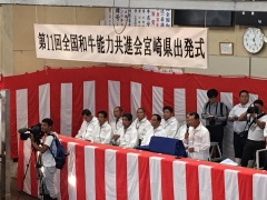 紅白幕で囲まれた場所に着席している、白い作業服を着た9名の男性の写真
