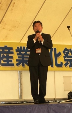 黒いスーツを着た市長が舞台上で両手でマイクを持ちながら話をしている写真