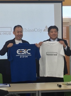 市長が白いTシャツを持っていて、隣で村岡さんが青いTシャツを持って笑顔で並んでいる写真
