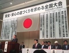 頭上に安心・安全の道づくりを求める全国大会と書かれた文字と日本国旗が掲げられていて、関係者達が座っている写真