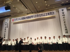 祝賀会にて舞台上に並ぶ白いジャケットを着た関係者の前で男性が何かを渡している写真