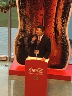 コカコーラのロゴが入った演台でマイクも持って話をしている市長の写真