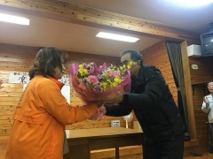 男性とオレンジのジャンパーを着た女性が向かい合って花束贈呈をしている写真