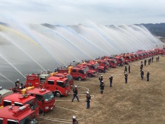 消防車が横にずらりと並んでいて、ホースから水が一斉に出ている写真