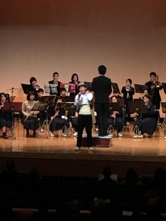 舞台上にマイクを持った少年、指揮者、吹奏楽の人たちが写っている写真