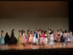 舞台上で色とりどりのドレスを着た女性たちがファッションショーのように歩いている写真
