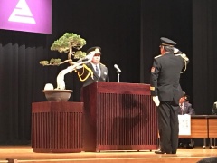 横に盆栽が置かれている舞台上で市長と関係者が向かい合って敬礼をしている写真