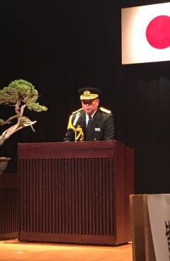奥に日本国旗が掲げられている舞台上でえびの市長が軍服のような制服を着て話をしている写真