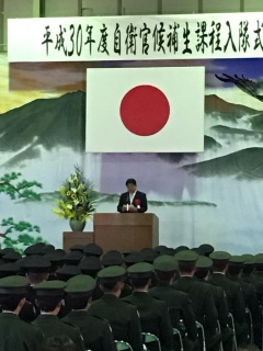 頭上に日の丸が掲げられた舞台でスーツを着た市長が話をしている写真