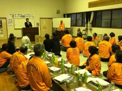 奥には話をする市長、手前にはオレンジの上着を着た参加者達が座っている写真