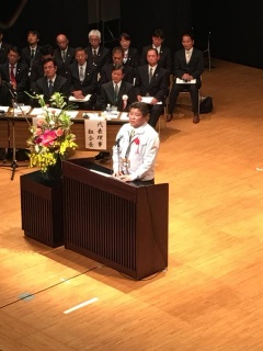 白い上着を着た市長が横に花が置かれている講演台で話をしている写真