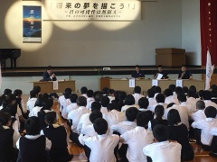 舞台前の議席に着席する市長や関係者と、体育座りをして並んでいる高校生の写真