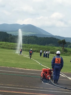 ゼッケン3を付けたヘルメット被った消防団員の先の芝生の上で放水活動をしている2名の消防団員と、消防の制服を着た数名の写真