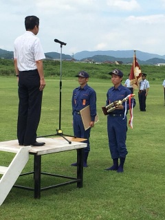 芝生の上でトロフィーと賞状を持っている二人の消防団員と壇上の上で立っている男性の写真