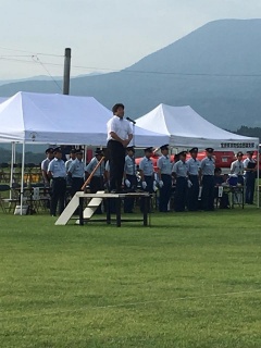 芝生の上で白いテントの前に整列し、多数の消防団員の前に置いてある壇上の上でマイクの前に立っている市長の写真