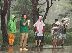 雨が降っている中、マイクを持っている男性と傘を差してる男性2名、雨衣を着ている2名の男性の写真