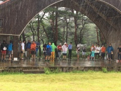 木でできた屋根のアーチ状のステージで雨宿りをしている大勢の参加者の写真