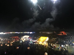 暗い夜空に煙を出しながら打ちあがる花火と、大勢の観客や出店の明かりが灯っている地上の写真