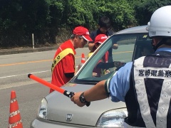 白いヘルメットを被った警察官が蛍光棒でグレーの自動車を停止させ、赤色の帽子と赤色の洋服を着た男女が運転手に話かけている写真