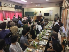 料理が置かれた長机に座っている多くの高齢者と前方の演台に立っている市長の写真