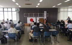 日本国旗がある会場前方方向に向かって並べられた机といすに座っている多くの参加者を後方から写した写真