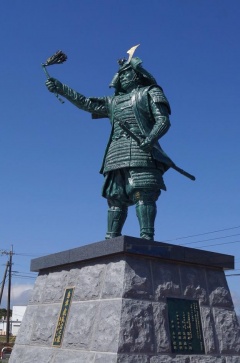 青空の下に立っている右手に旗、左手に日本刀をもって甲冑を着ている銅像の写真