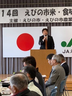 日本国旗が掲げられている舞台上で市長がマイクを持って話をしている写真