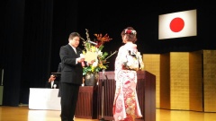 舞台上で、市長が振袖の女性の前で賞状を読み上げている写真