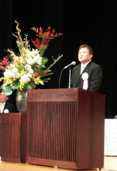 胸章を付けた市長が、花が飾られた演台に立っている写真
